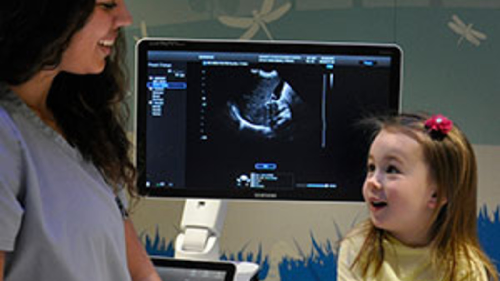 Sonographer chatting with a small child with an ultrasound on a computer monitor in the background